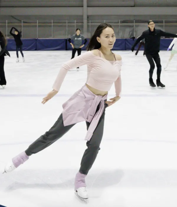 Young woman in dark pants and light pink ballet top ice skate on ice with other skaters behind her